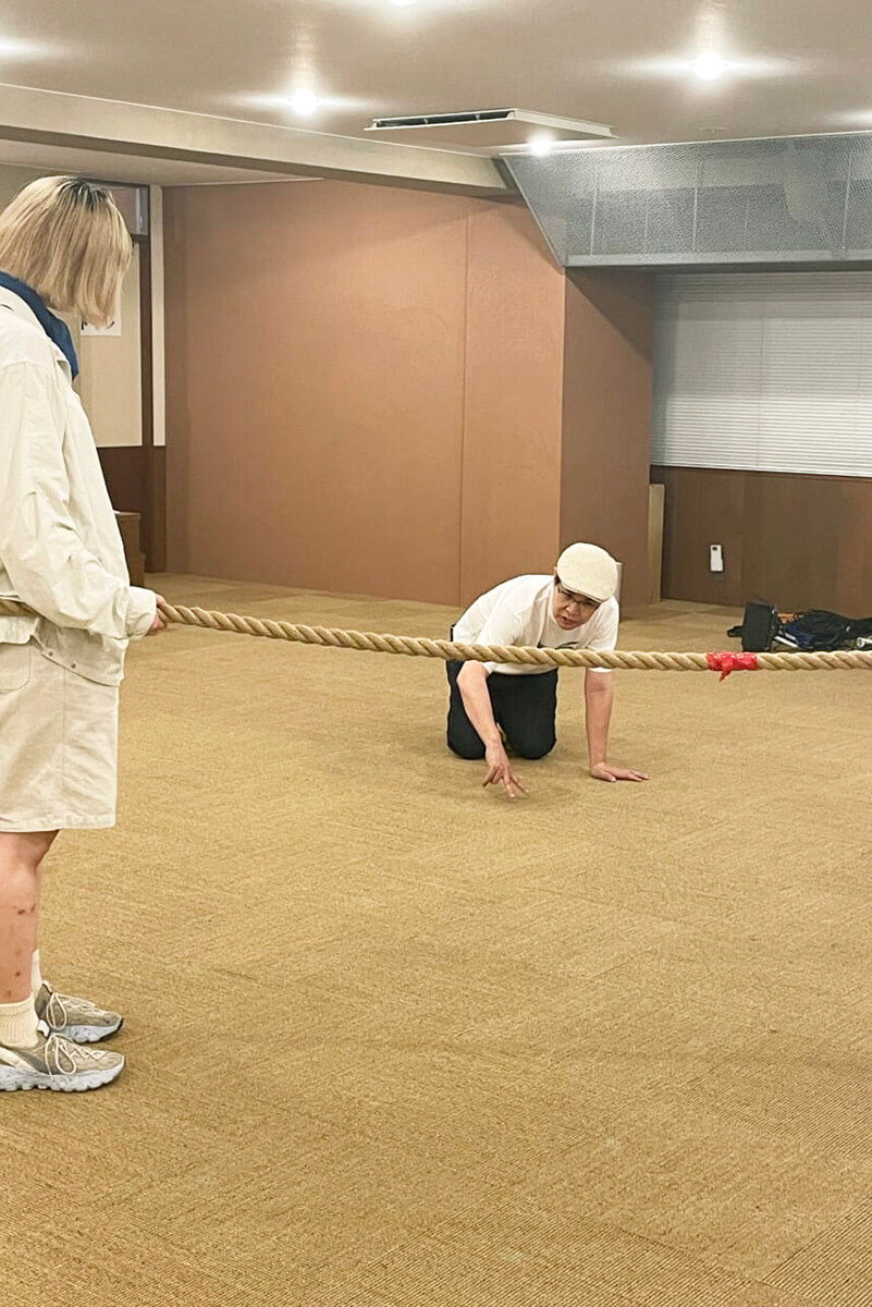 山下さんが膝をついて、指を地面につけてつなひきの姿勢を表しています。それを新メンバーのなつきさんが見て学んでいます。