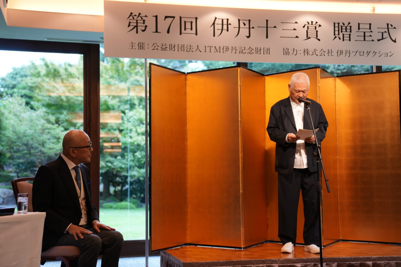 金の屏風の前で南伸坊さんが壇上に上がり、両手で紙を持って、マイクに向かって山田五郎さんへの祝辞を述べています。ステージの左側では、山田五郎さんが椅子に座って聞き入っています。