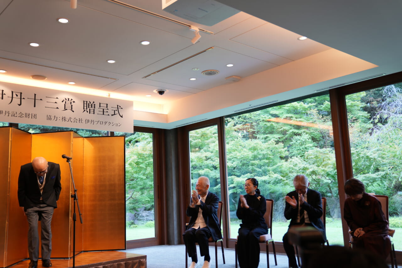 山田五郎さんが話し終わり、壇上でお辞儀をしています。ステージの右側で、選考委員の３名と宮本信子さんが並んで座り、拍手を贈っています。