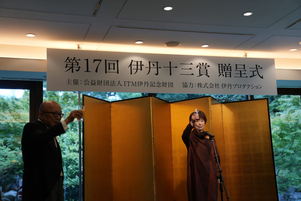 壇上で宮本信子さんがコップをかかげ、乾杯の合図をしています。左側では、山田五郎さんもコップをかかげています。