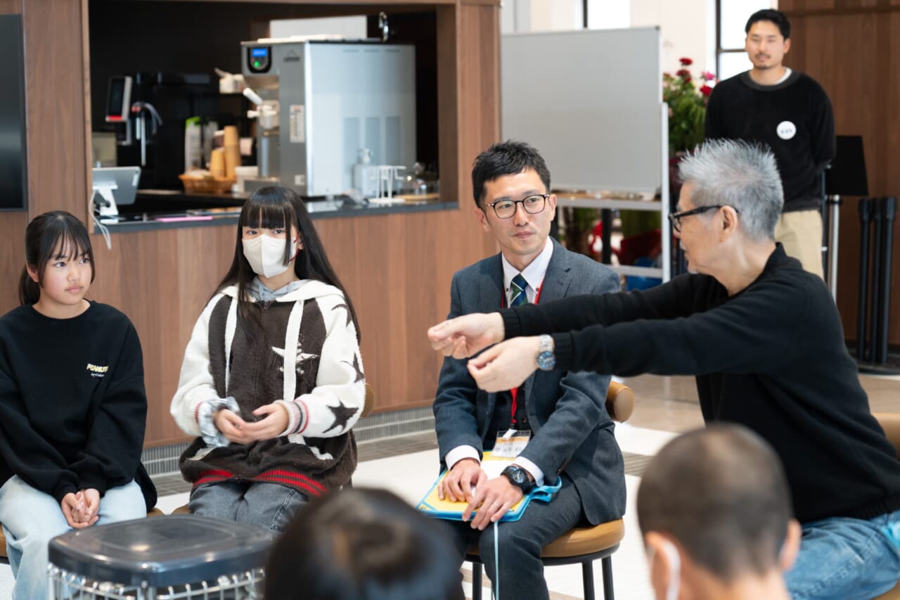 糸井がHさんのほうを向いて、ものを手渡すジェスチャーをしている。