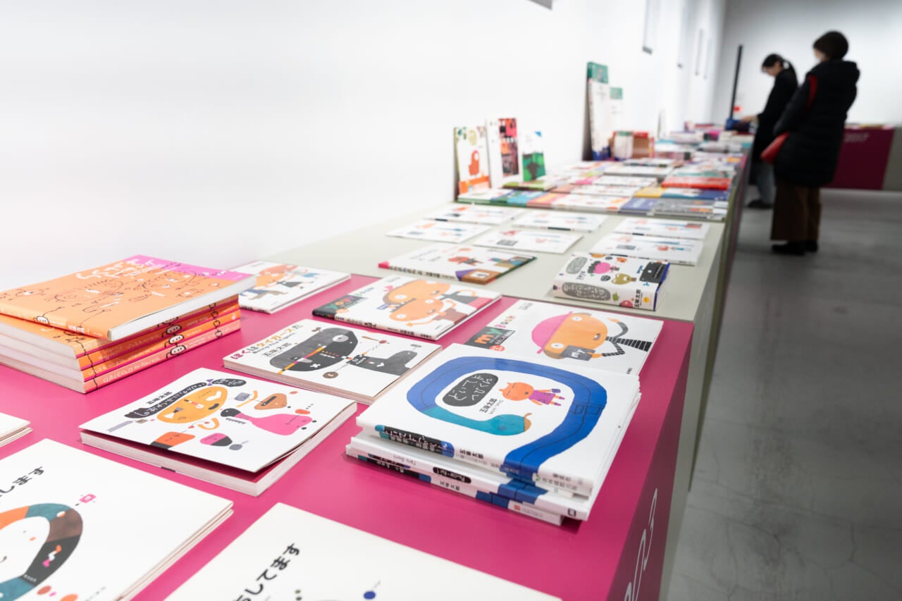 手前から奥に向かって、五味さんの絵本が机の上にずらっと並んでいる。奥には絵本や、壁にかかった五味さんの言葉を眺めているお客さんがいる。